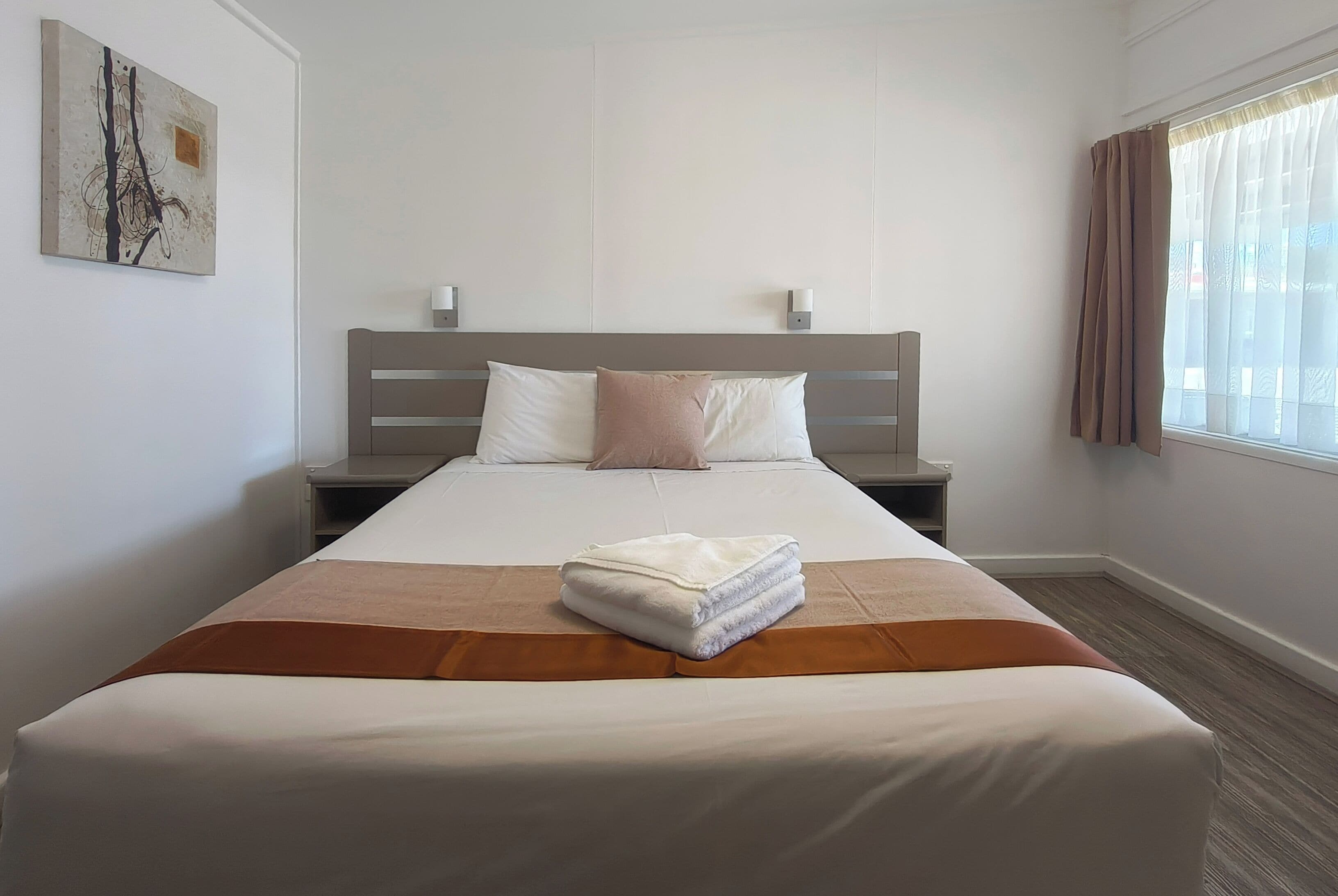 A hotel room with a bed, a headboard, two bedside tables, a wall-mounted artwork, and a window with curtains.