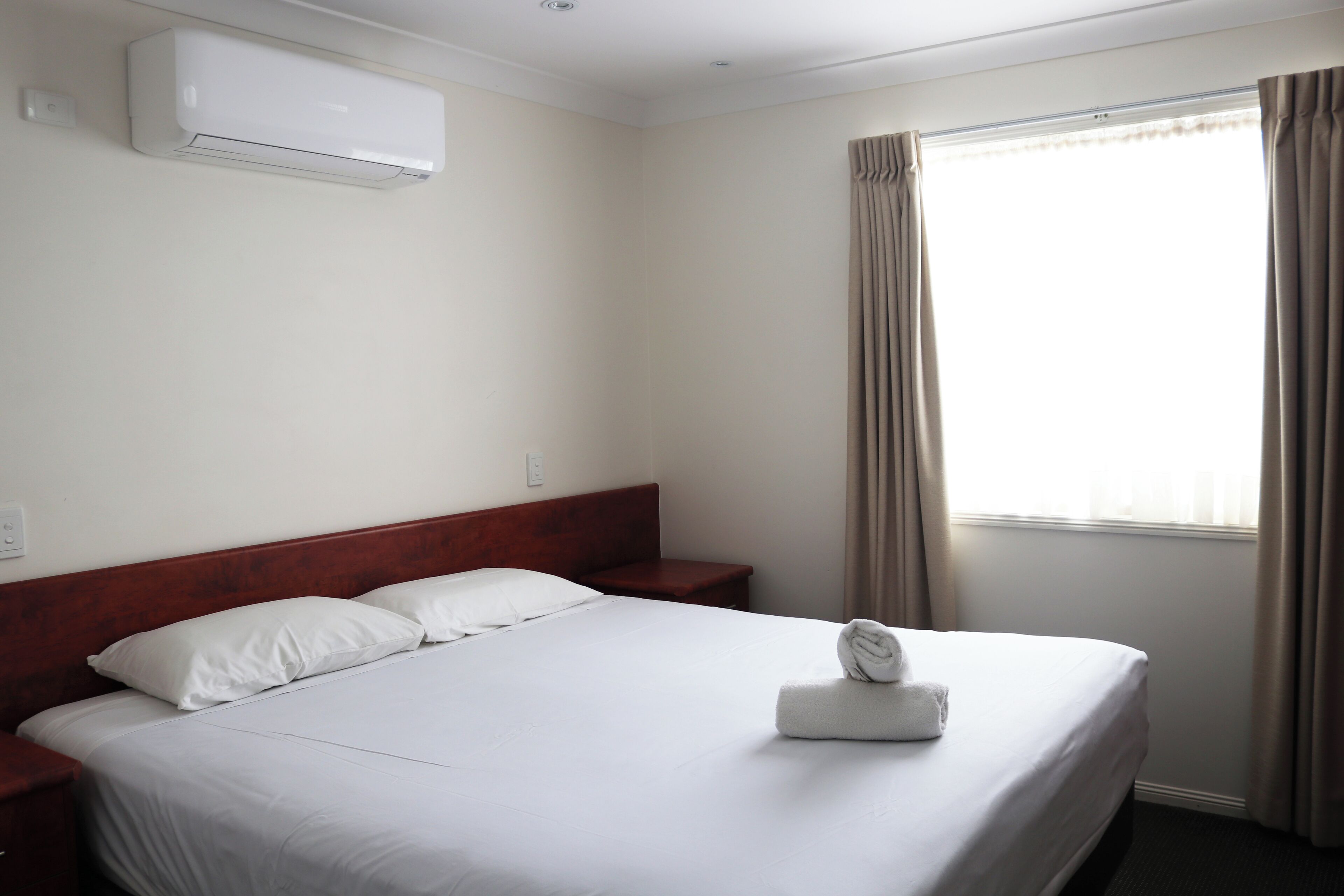 A hotel room with a bed, a nightstand, a wall-mounted air conditioner, and a window with curtains.