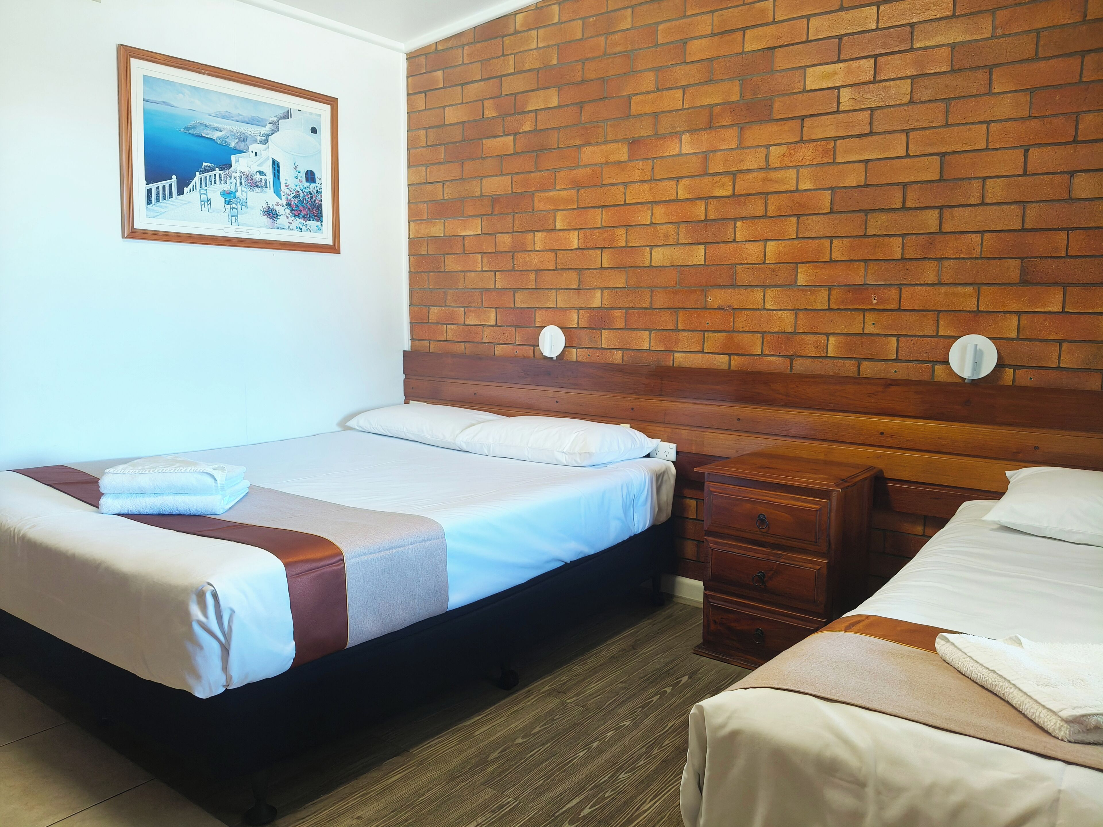 Hotellihuone, jossa on tiilikuviollinen sängynpääty, valkoisilla lakanoiden peitetty sänky, yöpöytä ja seinällä oleva kehystetty kuva.