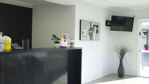 A reception area with a dark reception desk, a flat-screen TV mounted on the wall, a framed abstract painting, and a tall vase with long, thin plants. - Motel Myall (Dalby)
