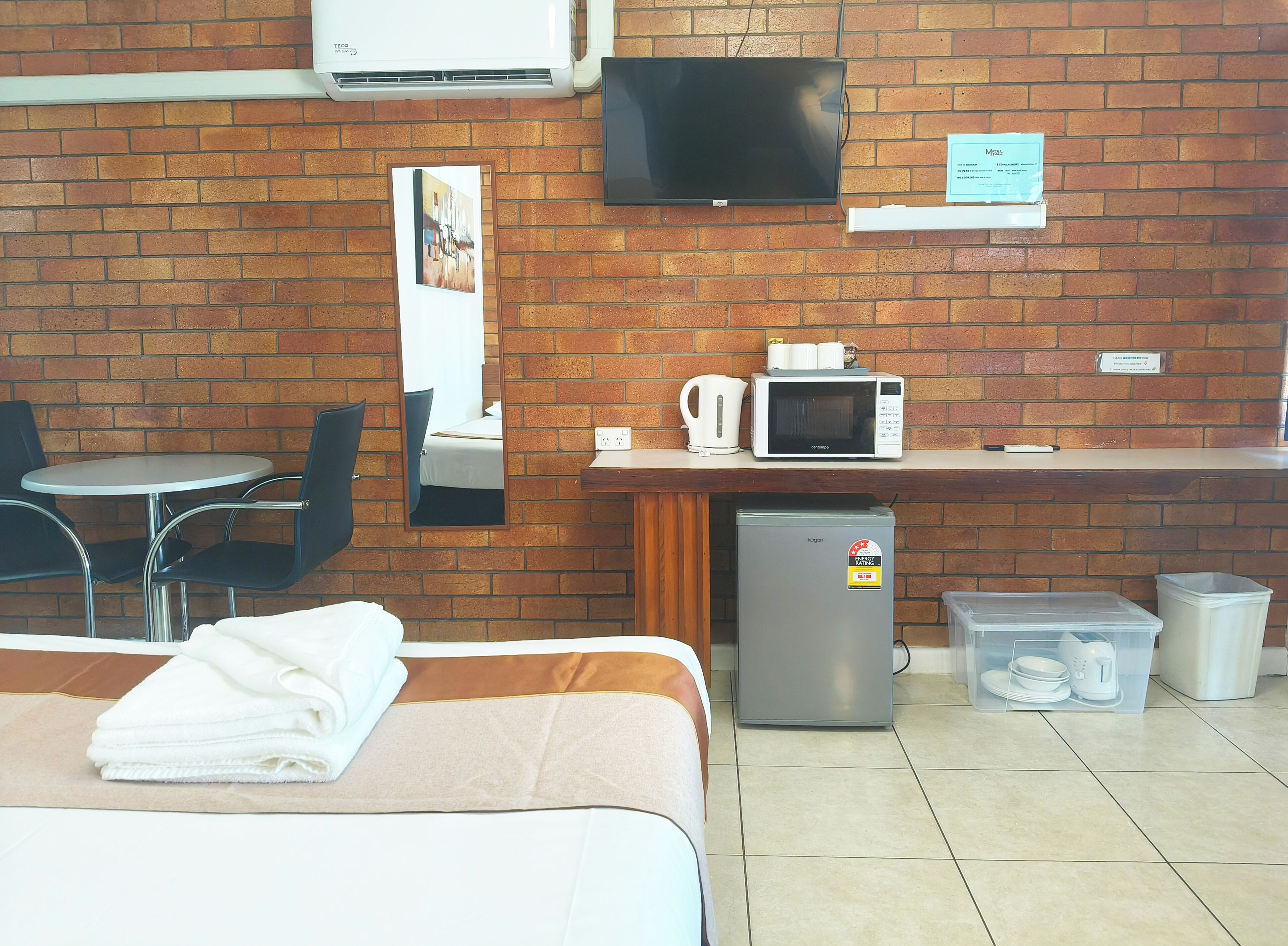 A hotel room with a bed, a desk with a microwave, a refrigerator, a TV mounted on the wall, and a brick wall.