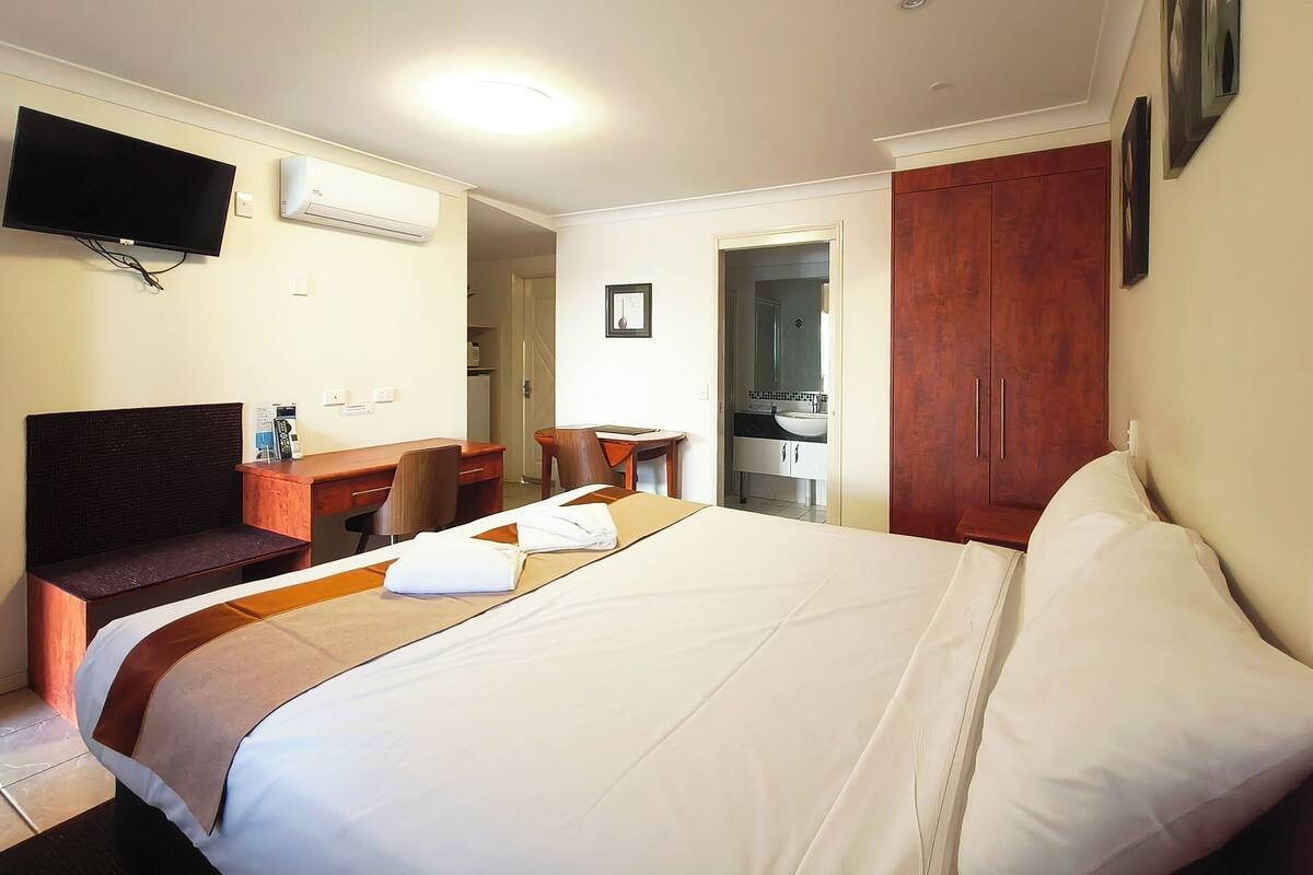 A hotel room with a bed, a desk, a chair, a TV, and an air conditioning unit.