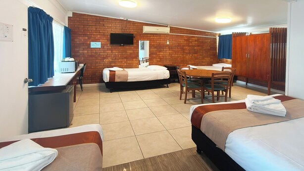 A hotel room with two beds, a dining table, chairs, a TV, and a brick wall. - Motel Myall (Dalby)