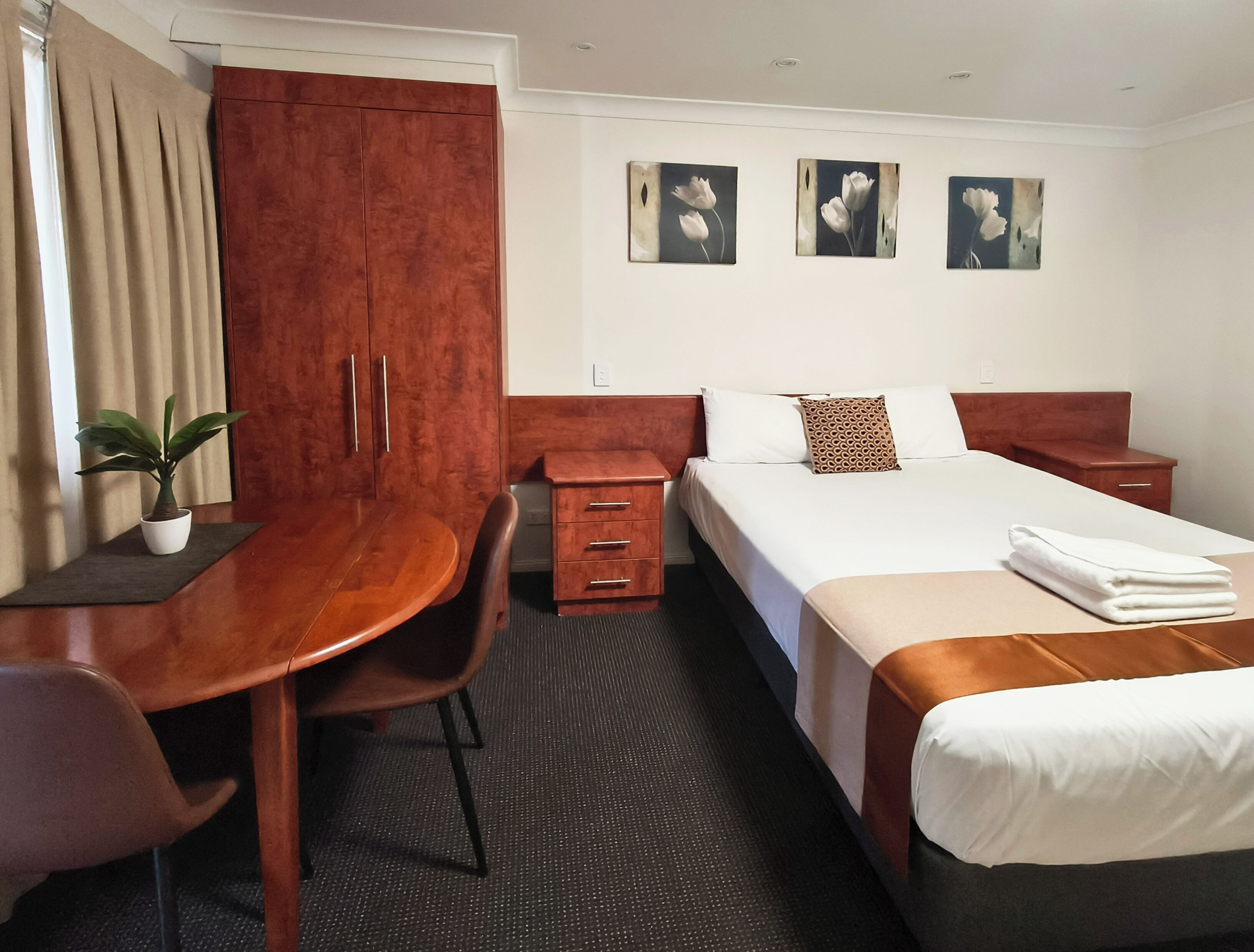 Una habitación de hotel con un escritorio de madera, una cama con ropa blanca, una mesita de noche, un armario y tres cuadros de flores colgados en la pared.