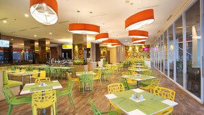 Een helder verlicht restaurant met groene en gele stoelen en tafels, grote ramen en een kleurrijk wandontwerp.