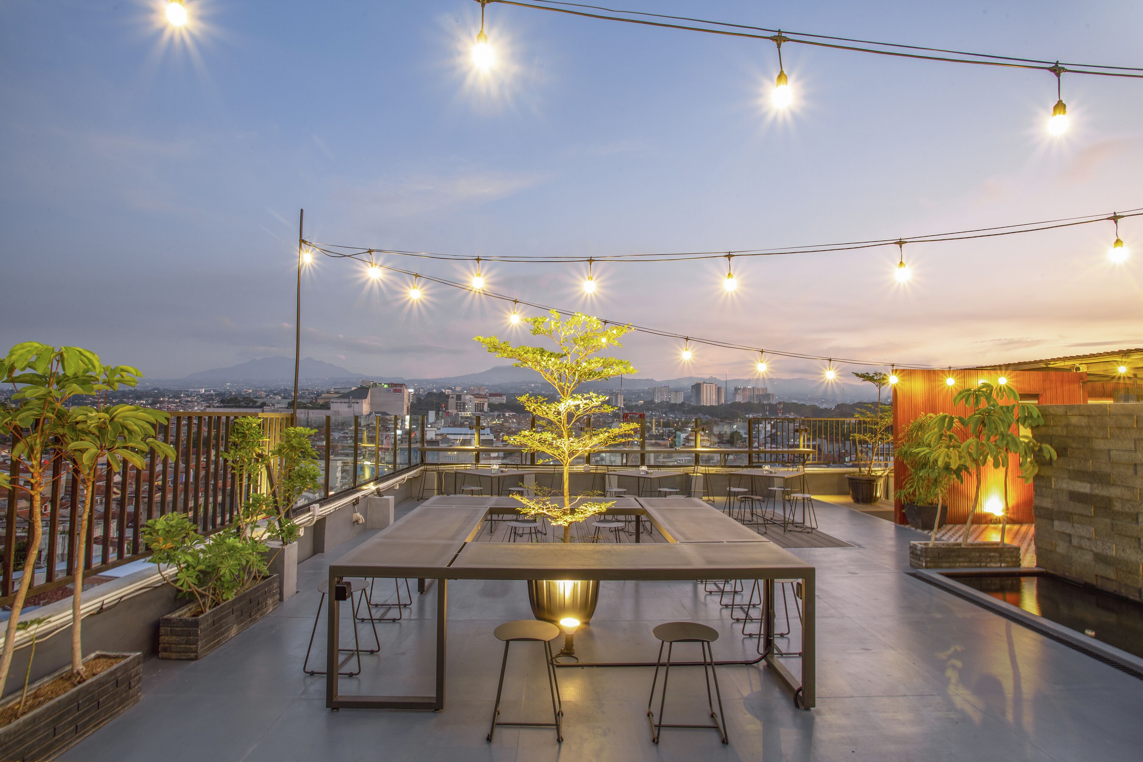 A rooftop terrace with a long dining table, bar stools, string lights, potted plants, and a view of the city and mountains.