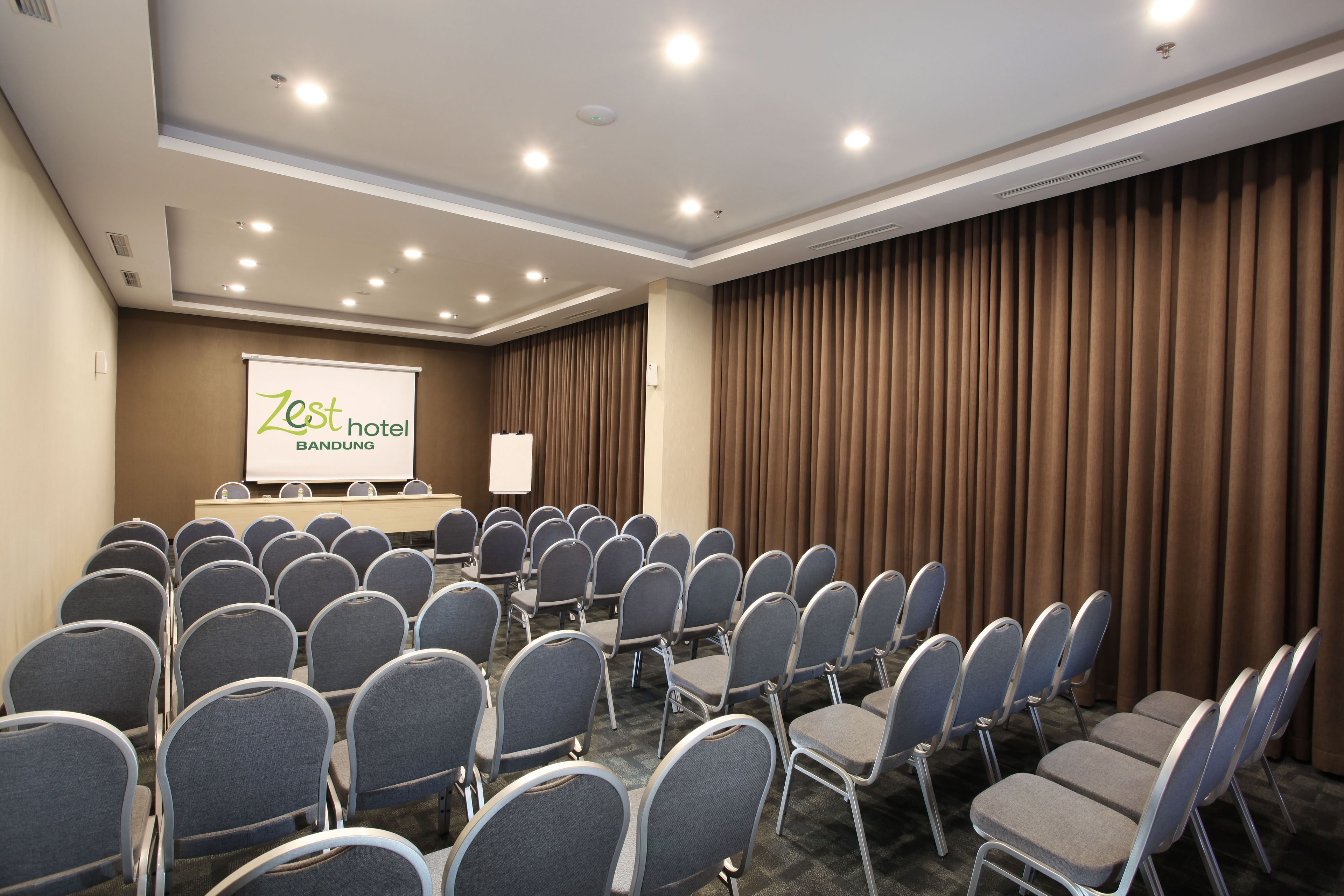 A conference room with rows of grey chairs, a podium, and a projector screen displaying "Zest Hotel Bandung".