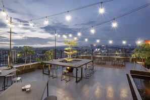 Een dakterras met tafels en stoelen, lichtjes en uitzicht op de stadsgezicht.