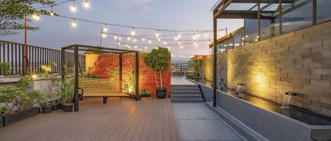 Una terraza en la azotea con luces colgantes, un banco de madera, un muro de ladrillo y una fuente de agua.