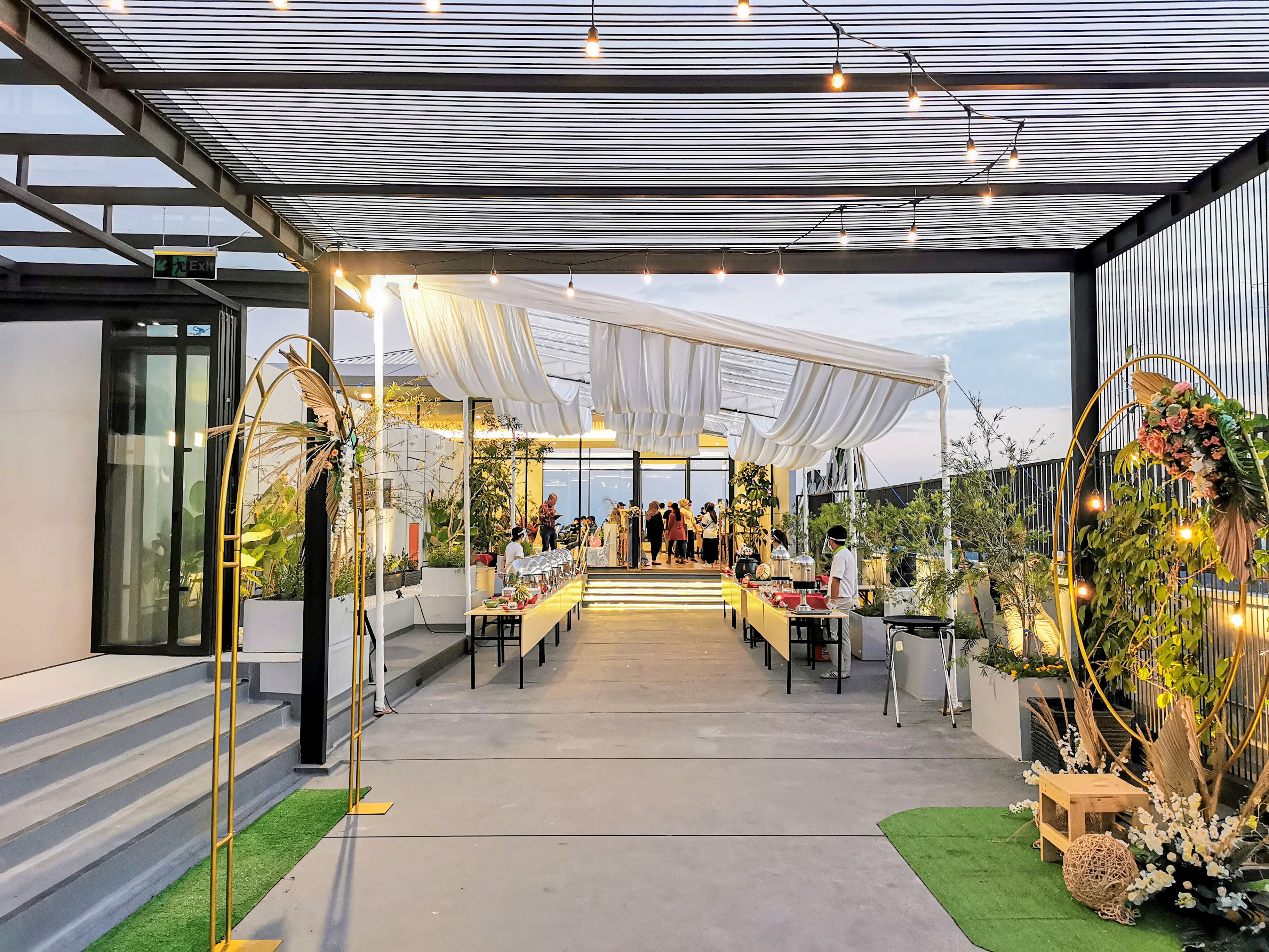 Un espacio interior para eventos con un pasillo cubierto, mesas dispuestas para comer y plantas en macetas.
