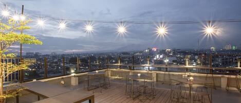 A rooftop terrace with tables and chairs, string lights, and a cityscape view.