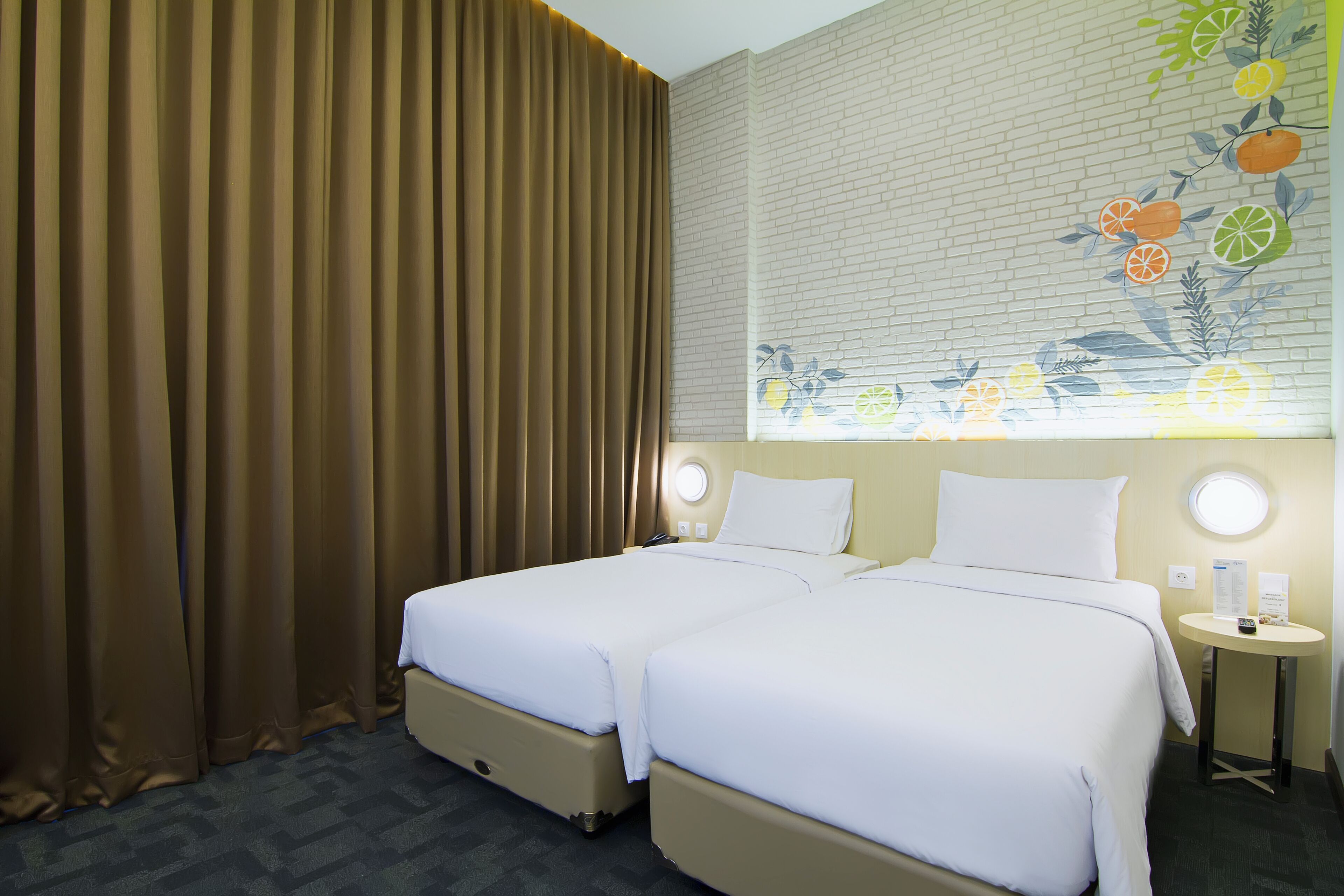Ein Hotelzimmer mit zwei Betten, einem Fenster mit einer Obstmalerei, einem runden Tisch mit einer Lampe und einem Vorhang.