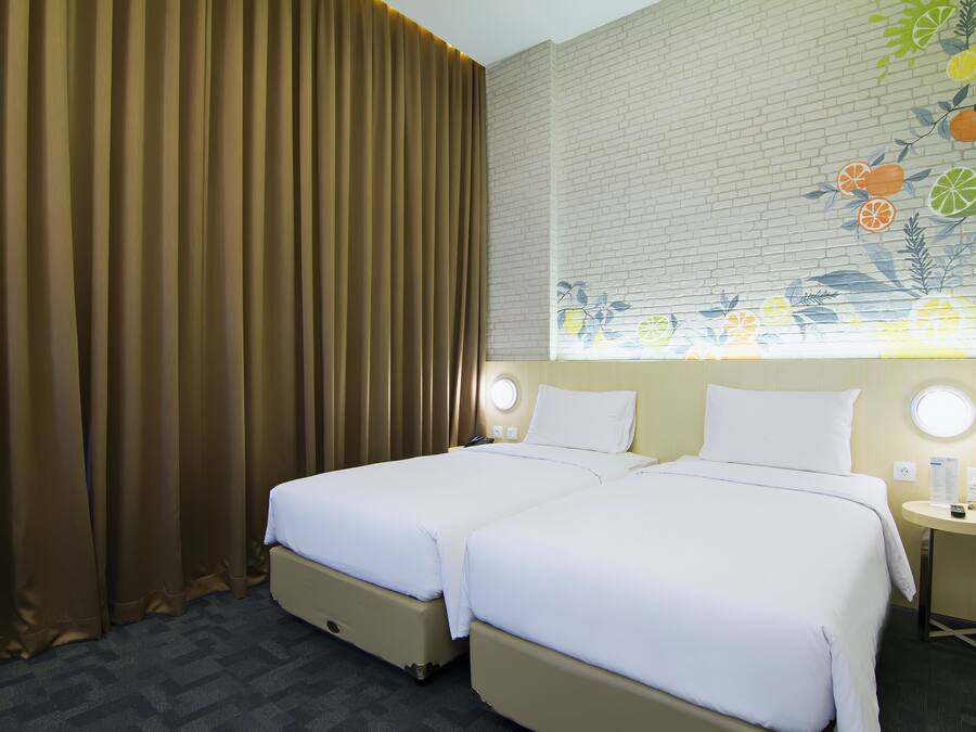 Ein Hotelzimmer mit zwei Betten, einem Fenster mit einer Obstmalerei, einem runden Tisch mit einer Lampe und einem Vorhang.