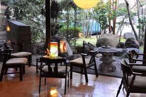 Un área de estar interior con mesas y sillas de piedra, una lámpara decorativa y vistas a un jardín con rocas y follaje.
