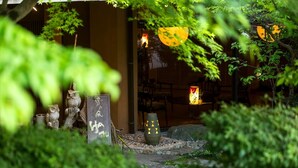 Un jardín japonés tradicional con faroles de piedra, un torii de madera y una casa de té visible entre el follaje.