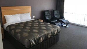 A hotel room with a bed, two black massage chairs, a nightstand, and a large window with curtains.