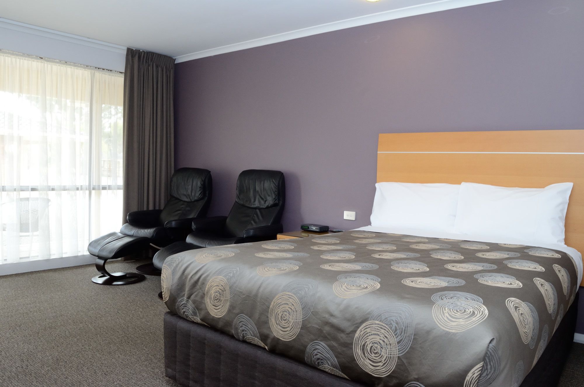 Habitación de hotel con una cama grande, dos sillones de cuero negro, un ventanal con cortinas y vistas al exterior.