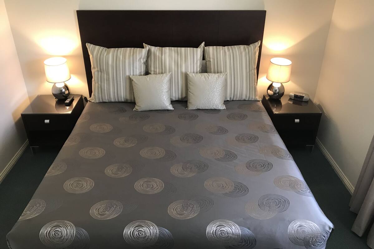 A neatly made bed with a patterned bedspread, two bedside lamps, and a dark headboard.