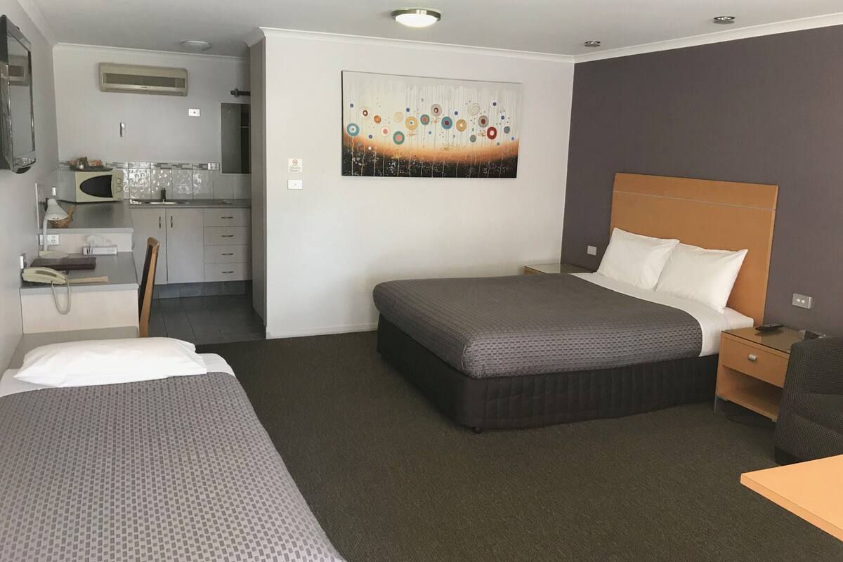 A hotel room with a bed, a desk, a chair, a small kitchenette, and a painting on the wall.