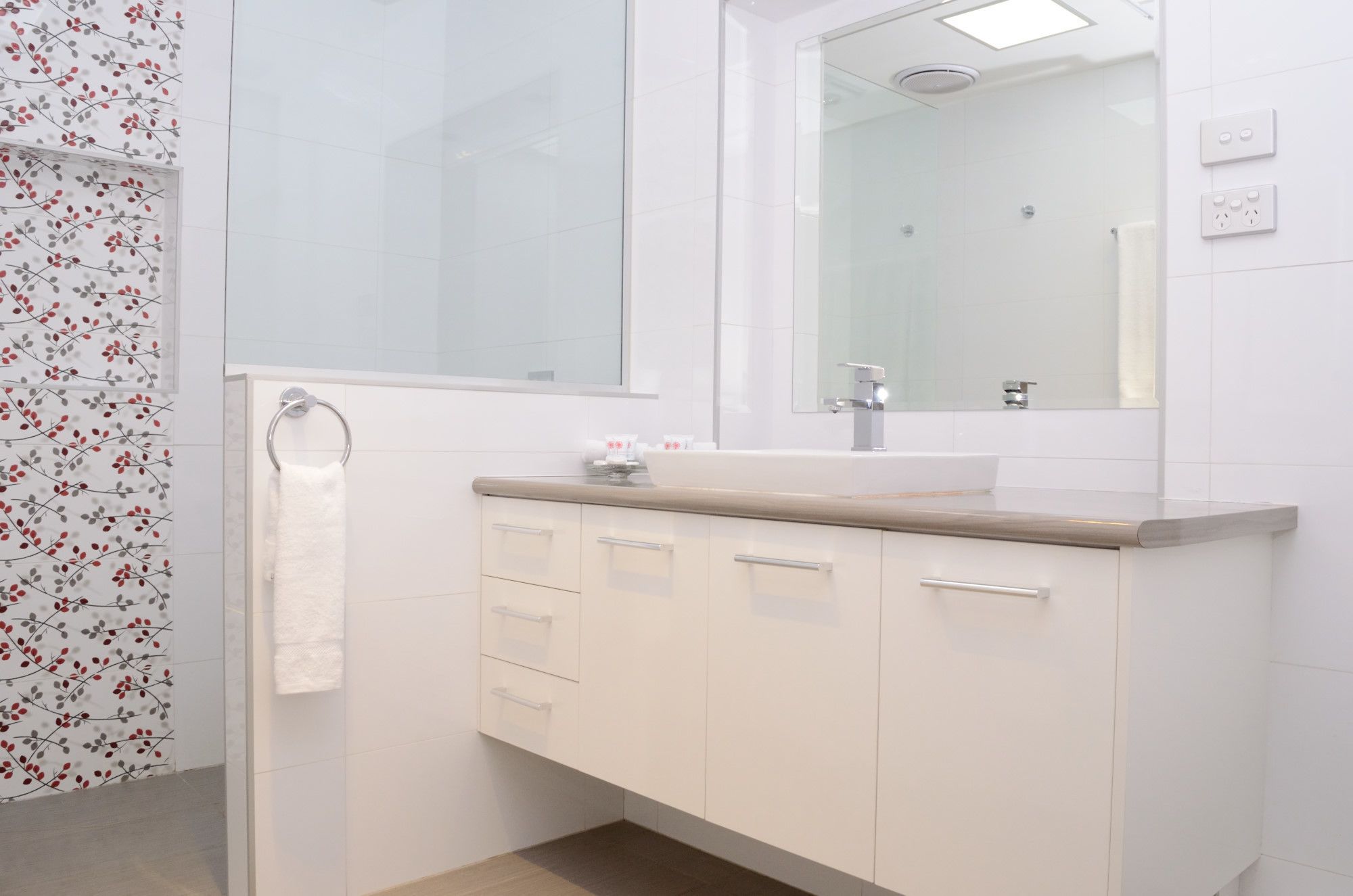 Un bany modern amb una cabina de dutxa de vidre, un lavabo blanc i una superfície de taulell beix, i un mirall sobre el lavabo.