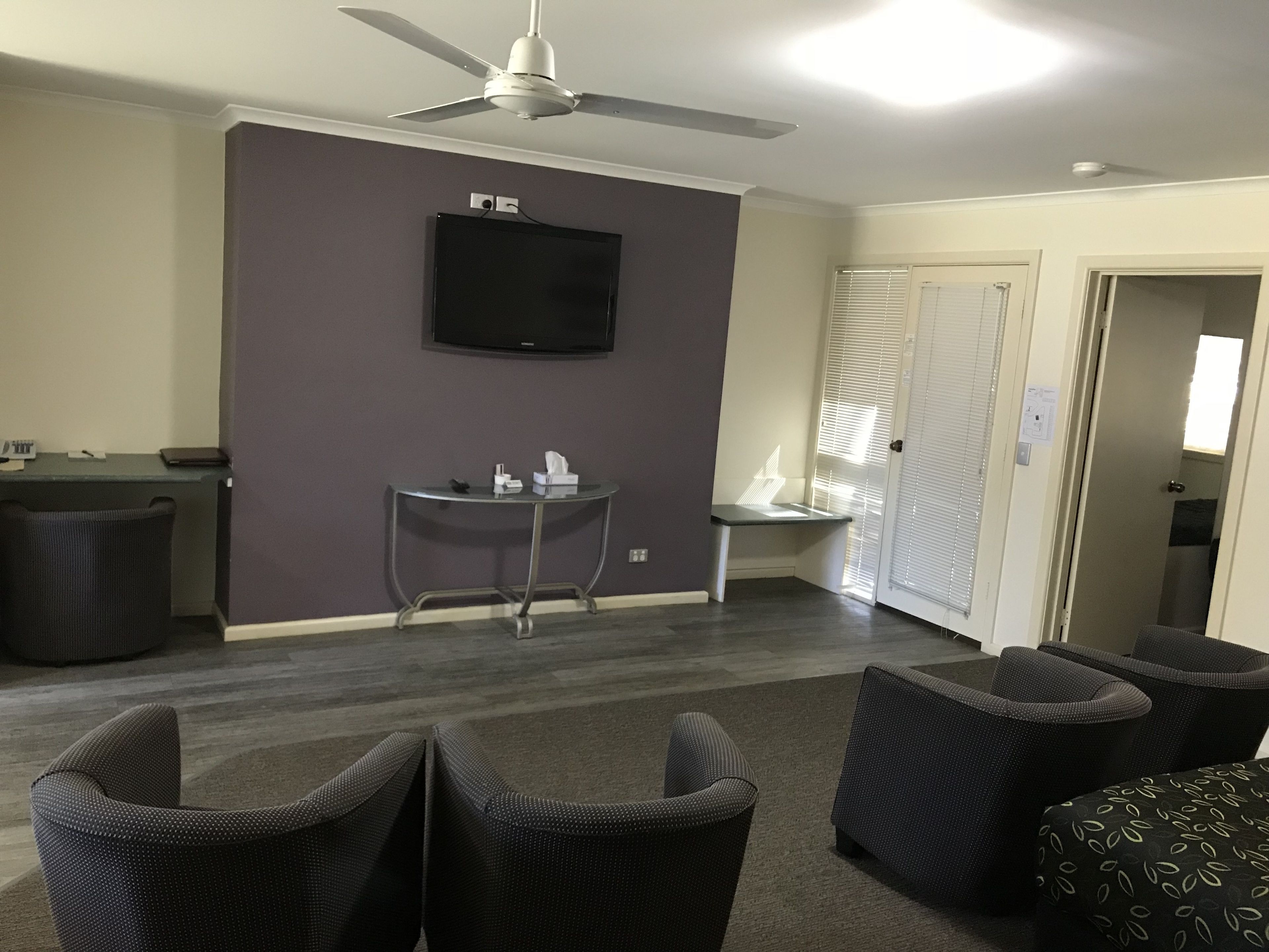 Un salon moderne avec un téléviseur à écran plat fixé au mur, un ventilateur de plafond et une petite table d’appoint sur laquelle se trouvent une boîte de mouchoirs et une télécommande. On y trouve quatre chaises de couleur foncée et un pouf à motifs.