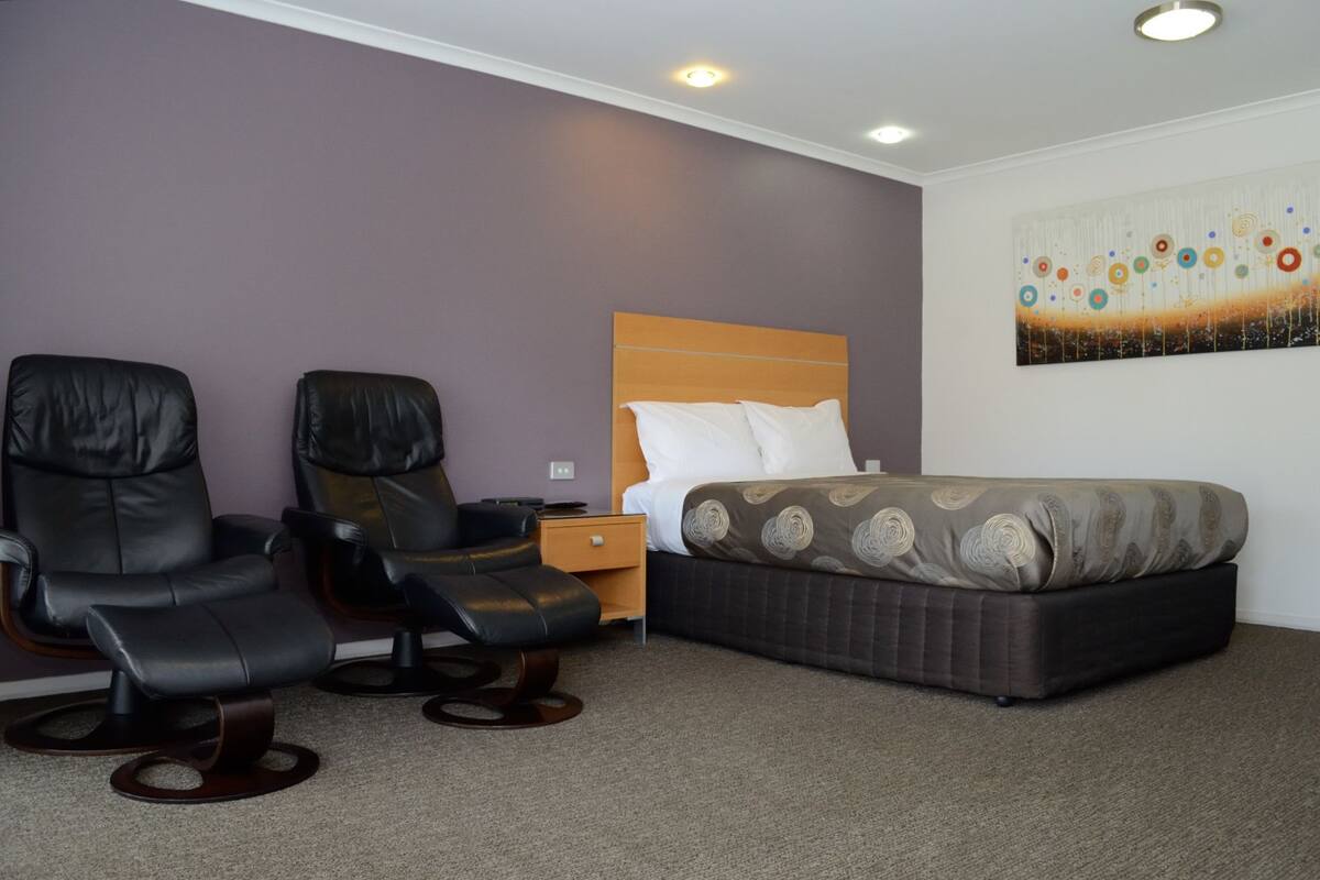 A hotel room with a bed, two leather chairs, a nightstand, and a wall art piece.