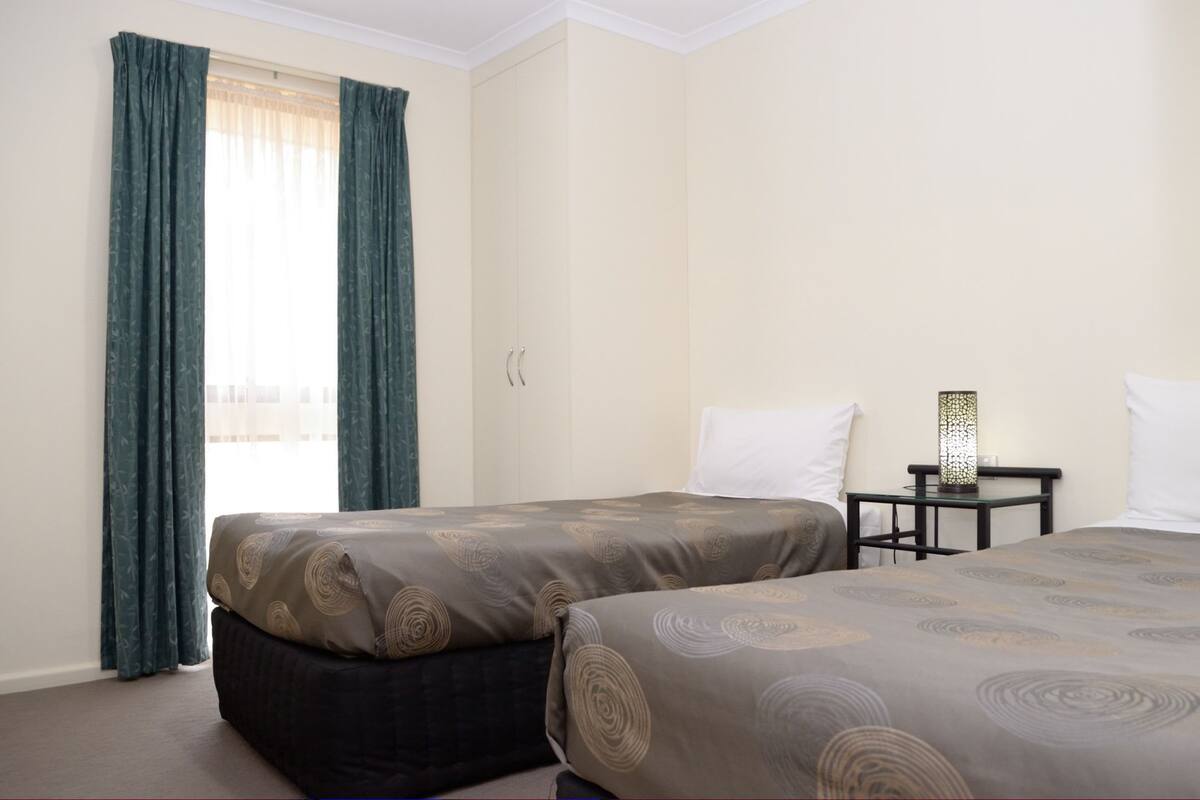 A hotel room with two beds, a nightstand, a lamp, and a window with curtains.