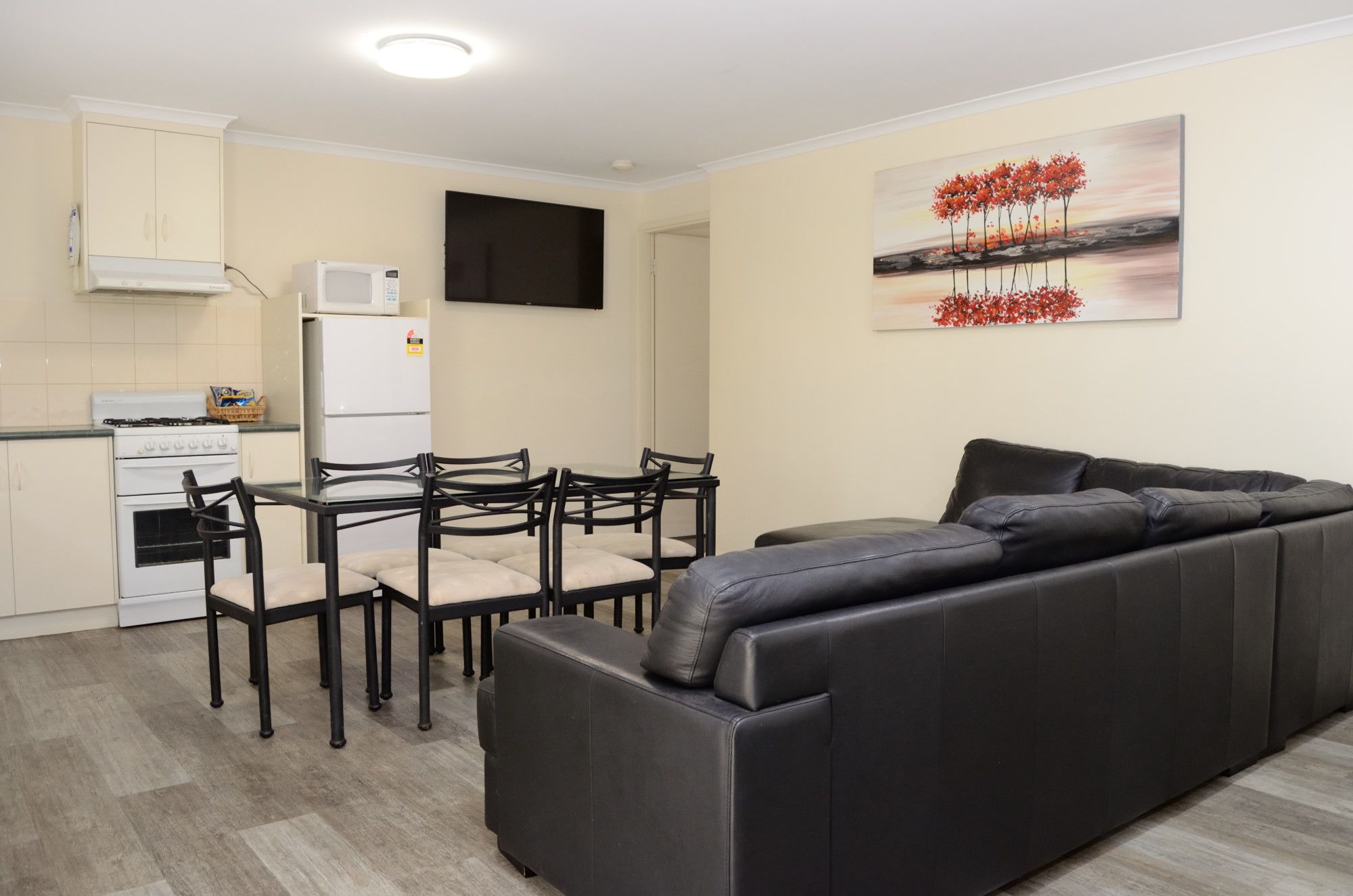 A modern living room with a black leather sofa, a dining table with chairs, a kitchen area with a stove and refrigerator, and a painting on the wall.