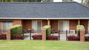 A two-story brick building with a dark tiled roof, a covered porch with a black metal fence, and red flowering bushes.