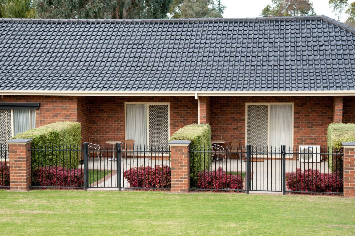 A two-story brick building with a dark tiled roof, a covered porch with a black metal fence, and red flowering bushes.