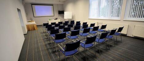 Un salón de conferencias con filas de sillas, una pantalla de proyector, una pizarra y una ventana con persianas.