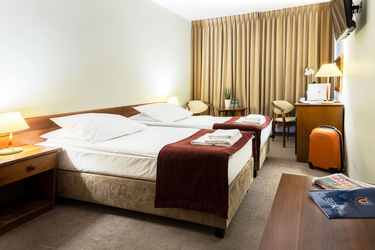 Una habitació d'hotel amb dos llits, una taula de treball, una cadira, una maleta i una làmpada.