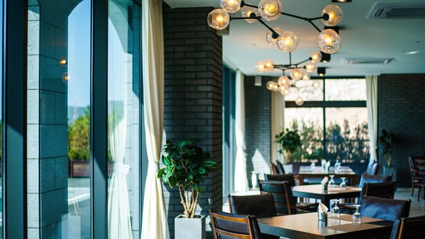 A restaurant interior with large windows, brick walls, and a modern chandelier. Tables are set with chairs, and there are potted plants. - Lapis Hotel (Namhae)