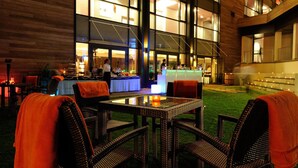 Un área de comedor al aire libre con sillas de mimbre y una mesa de vidrio, con vistas a un bar con mostrador y taburetes.