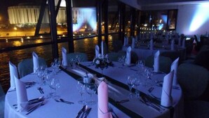 Comedor con mesas dispuestas para una comida, con vistas a un cuerpo de agua y un edificio al fondo.