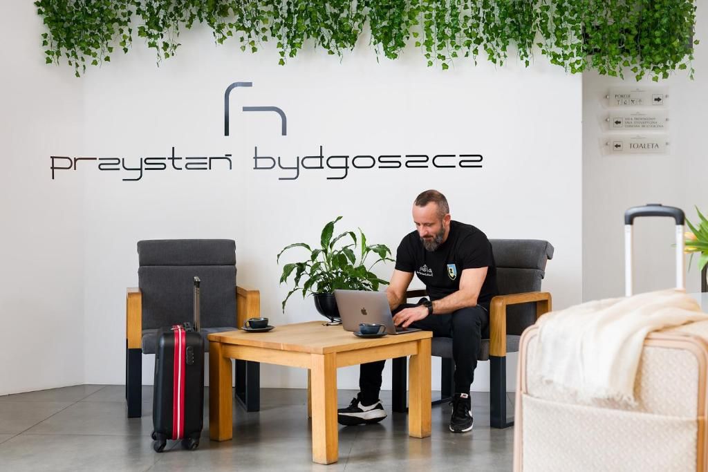 Een man zit aan een houten tafel met een laptop, een koffer en een plant in een kamer met een witte muur en een hangplant.