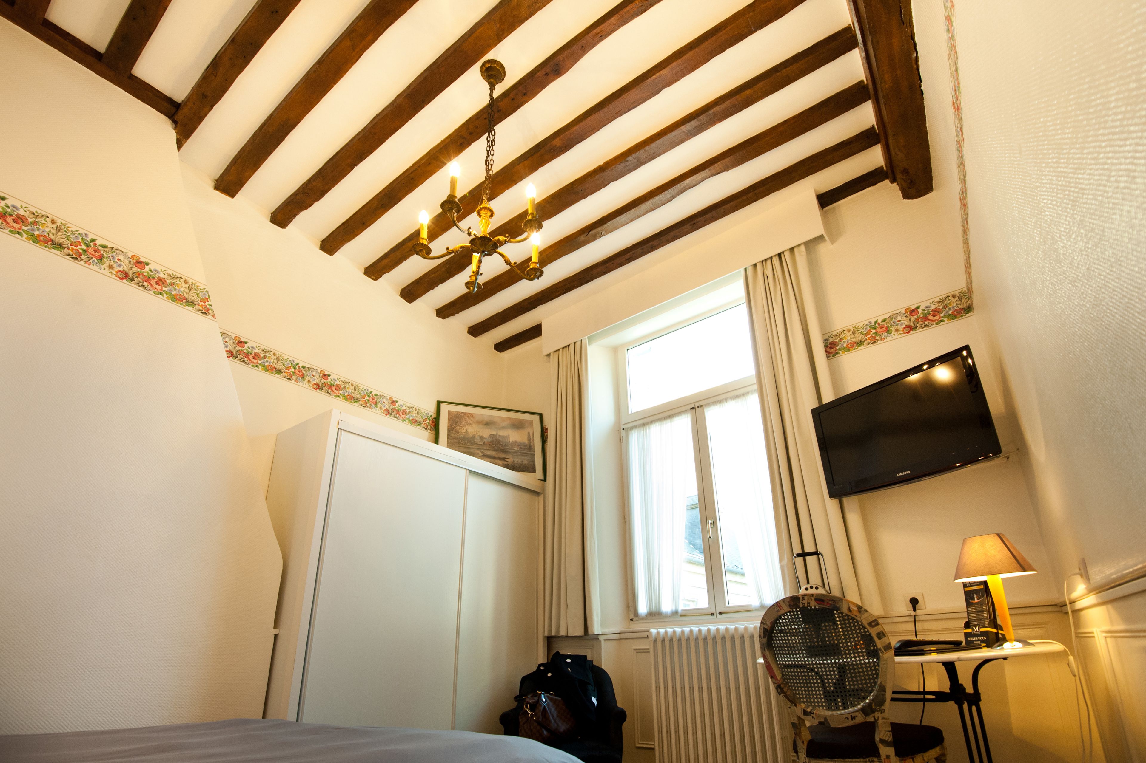 A hotel room with a wooden ceiling, a bed, a desk with a lamp, a TV, and a window with curtains.