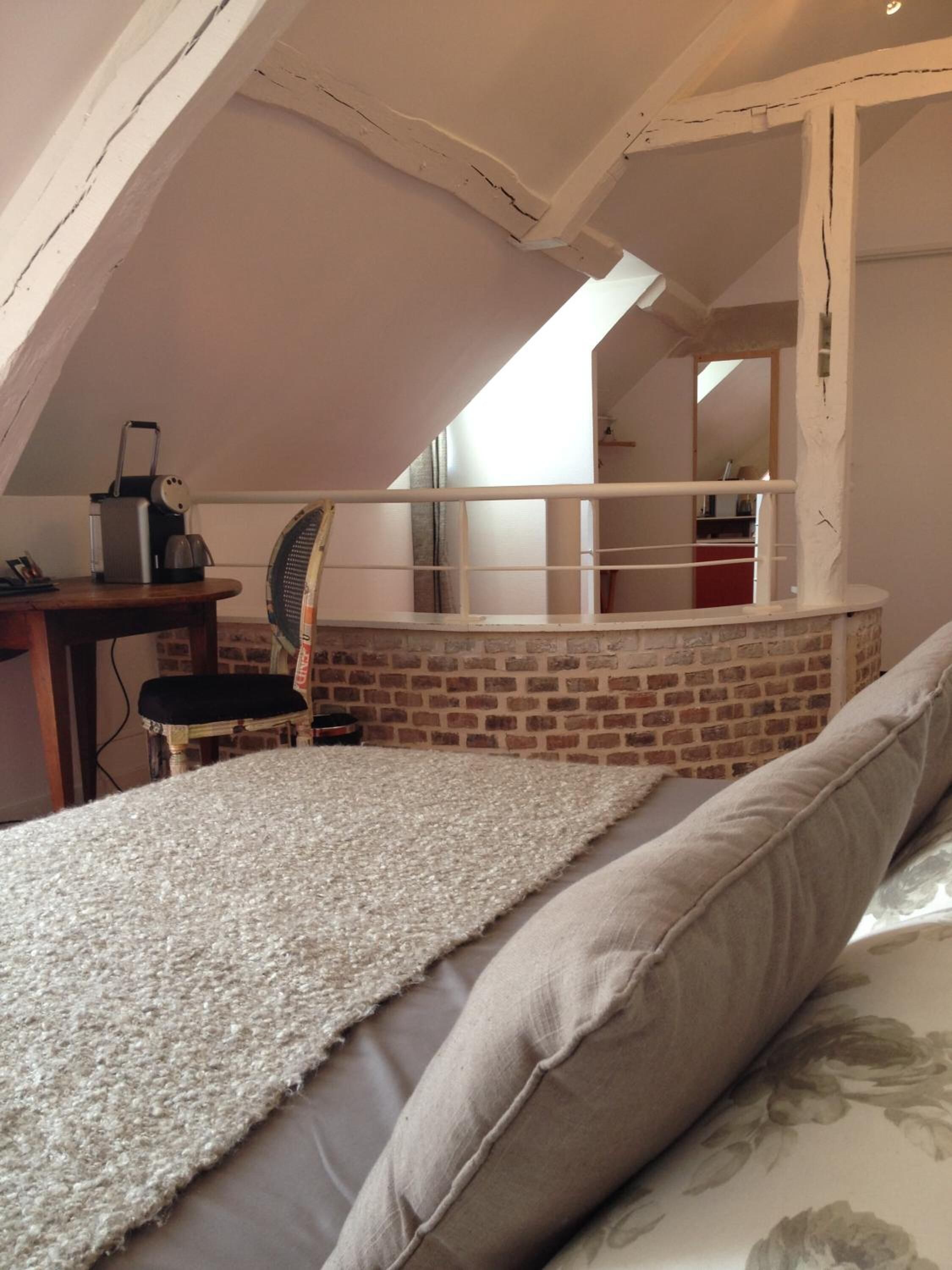 A bedroom with a sloped ceiling, a brick wall, a bed with a white textured blanket, a chair, and a desk with a lamp and a small heater.