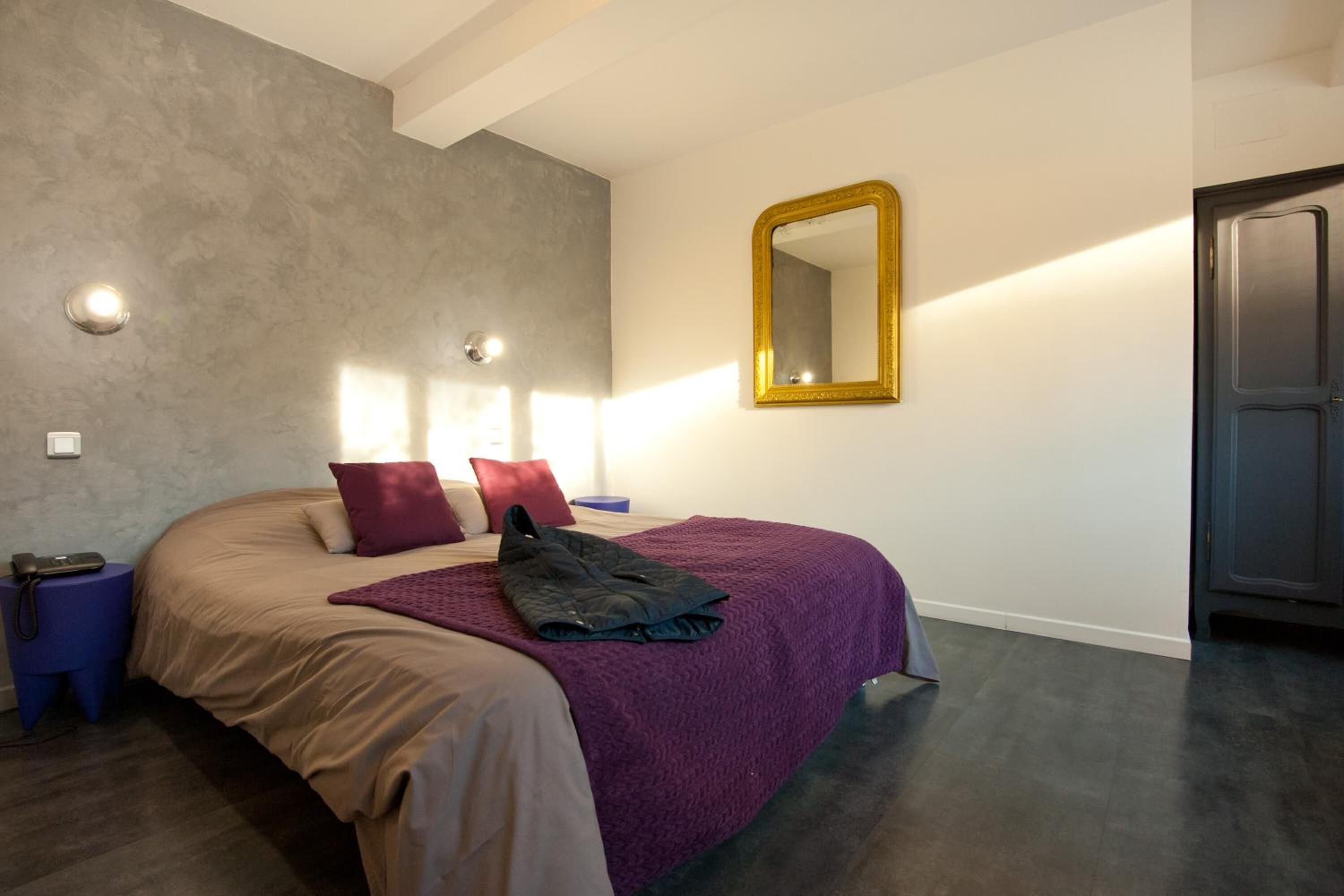 A bedroom with a bed, a purple blanket, a gold-framed mirror, and a door.