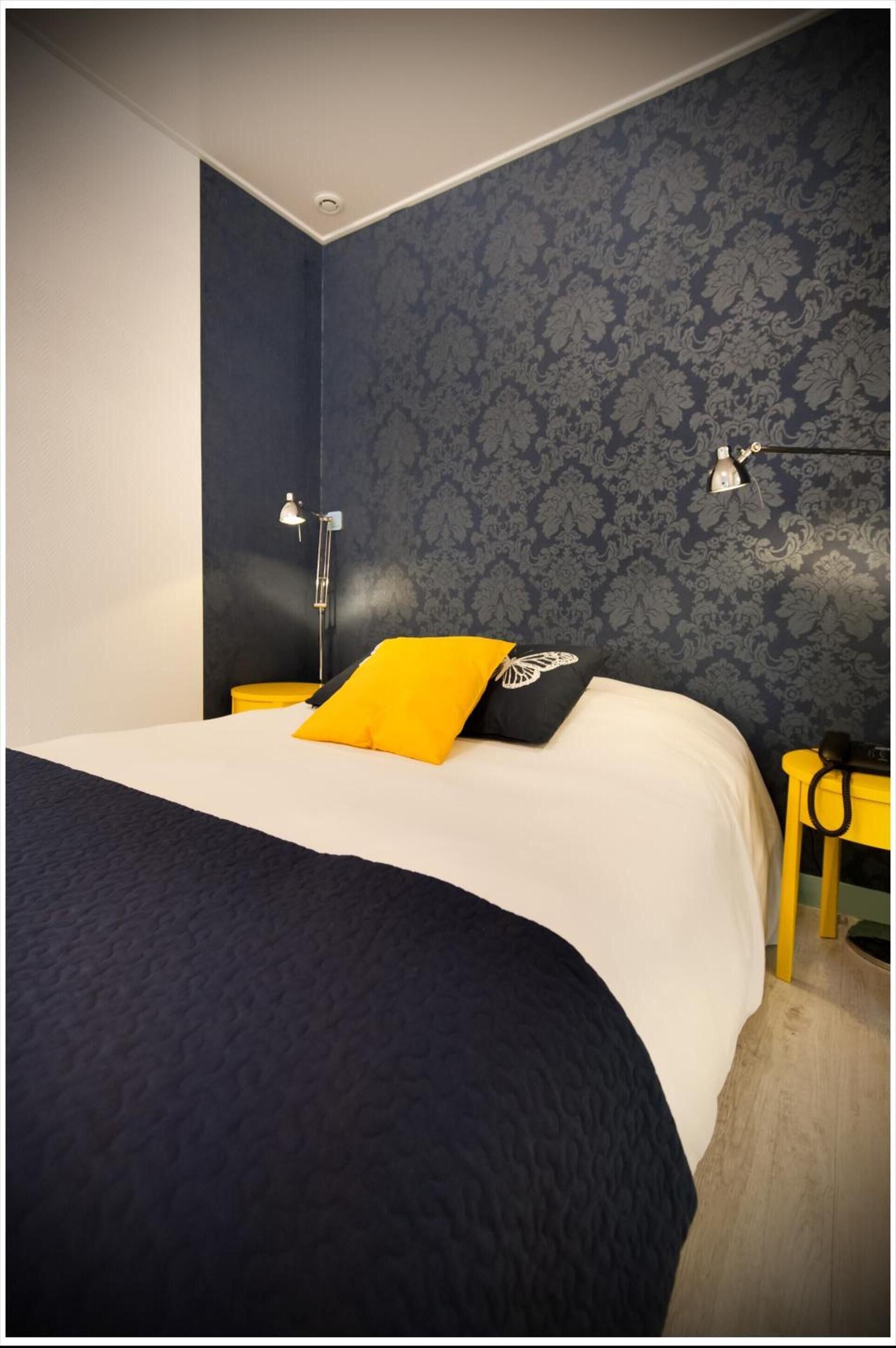 Quarto com uma cama grande, papel de parede azul escuro, um travesseiro amarelo e uma luminária de cabeceira.