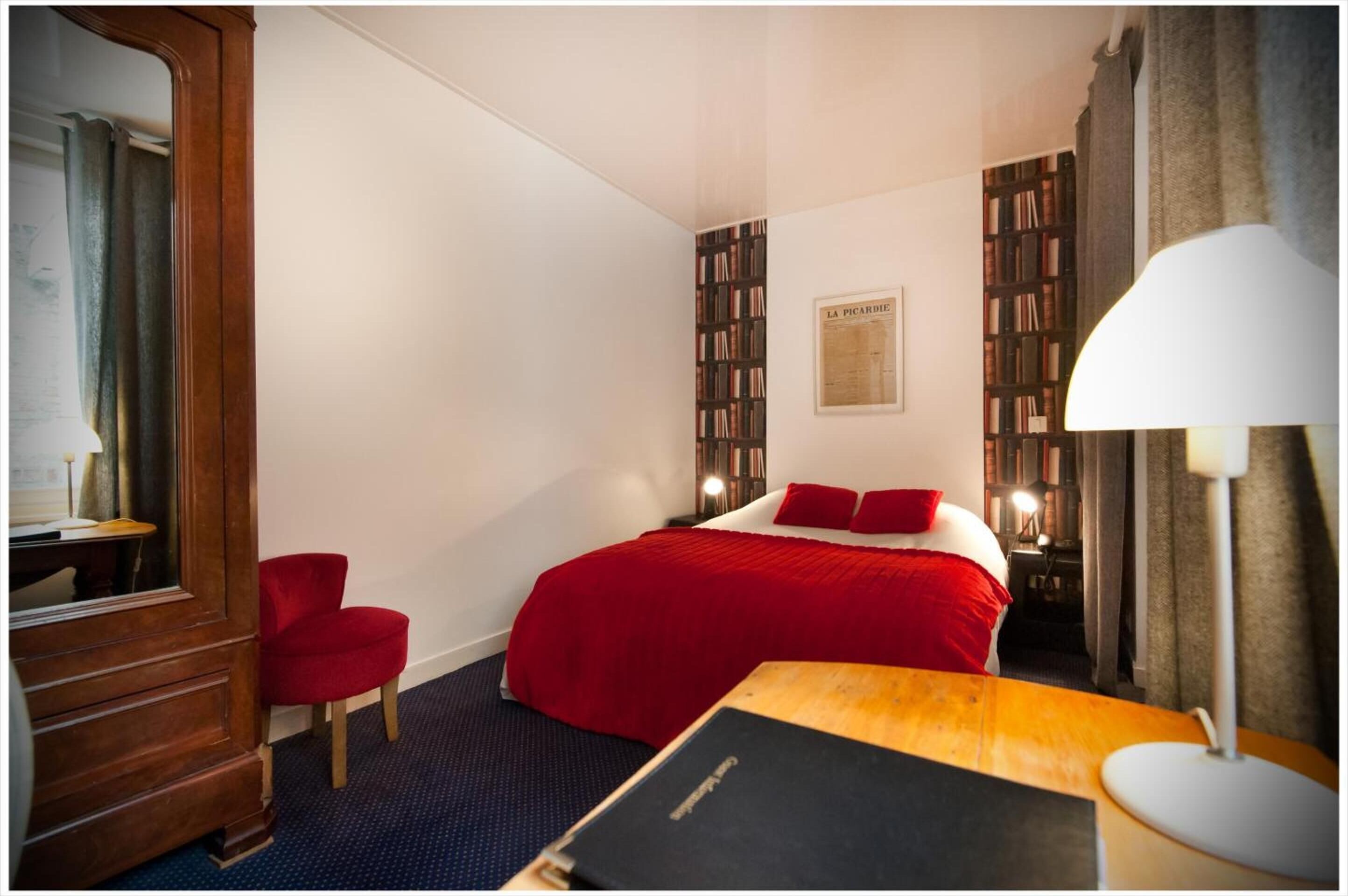 A hotel room with a large bed, a red chair, a wooden desk, a bookshelf, and a framed picture on the wall.
