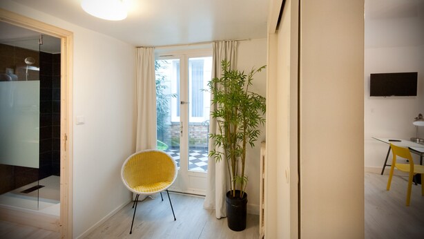 A modern room with a glass door leading to a balcony, a potted bamboo plant, a yellow chair, and a flat-screen TV mounted on the wall. - La Résidence le Prieuré (Amiens)