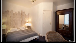 A bedroom with a bed, bedside lamps, a wooden headboard, a mirror, and a wicker chair. - La Résidence le Prieuré (Amiens)