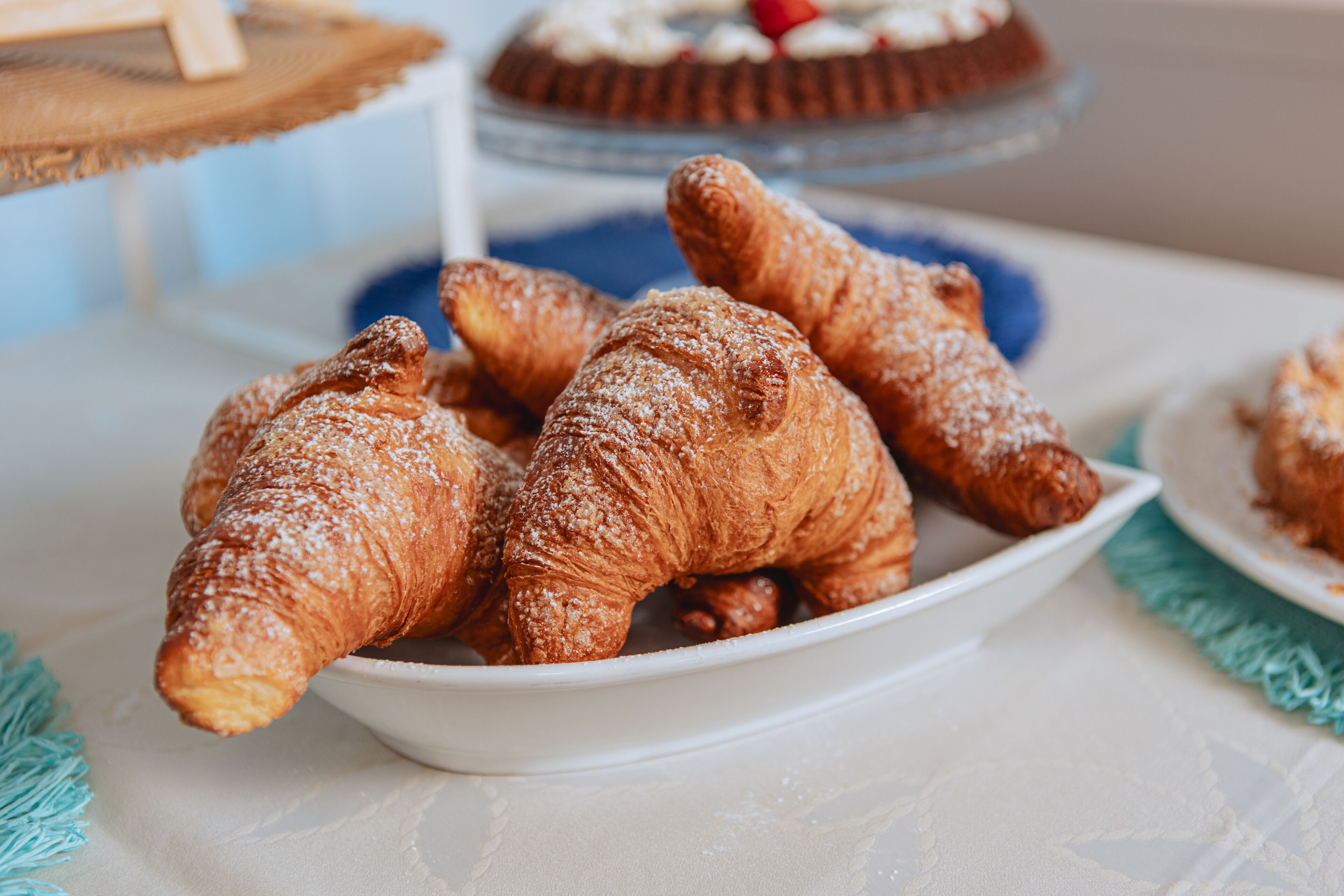 A white table with a plate of croissants and a cake in the background.