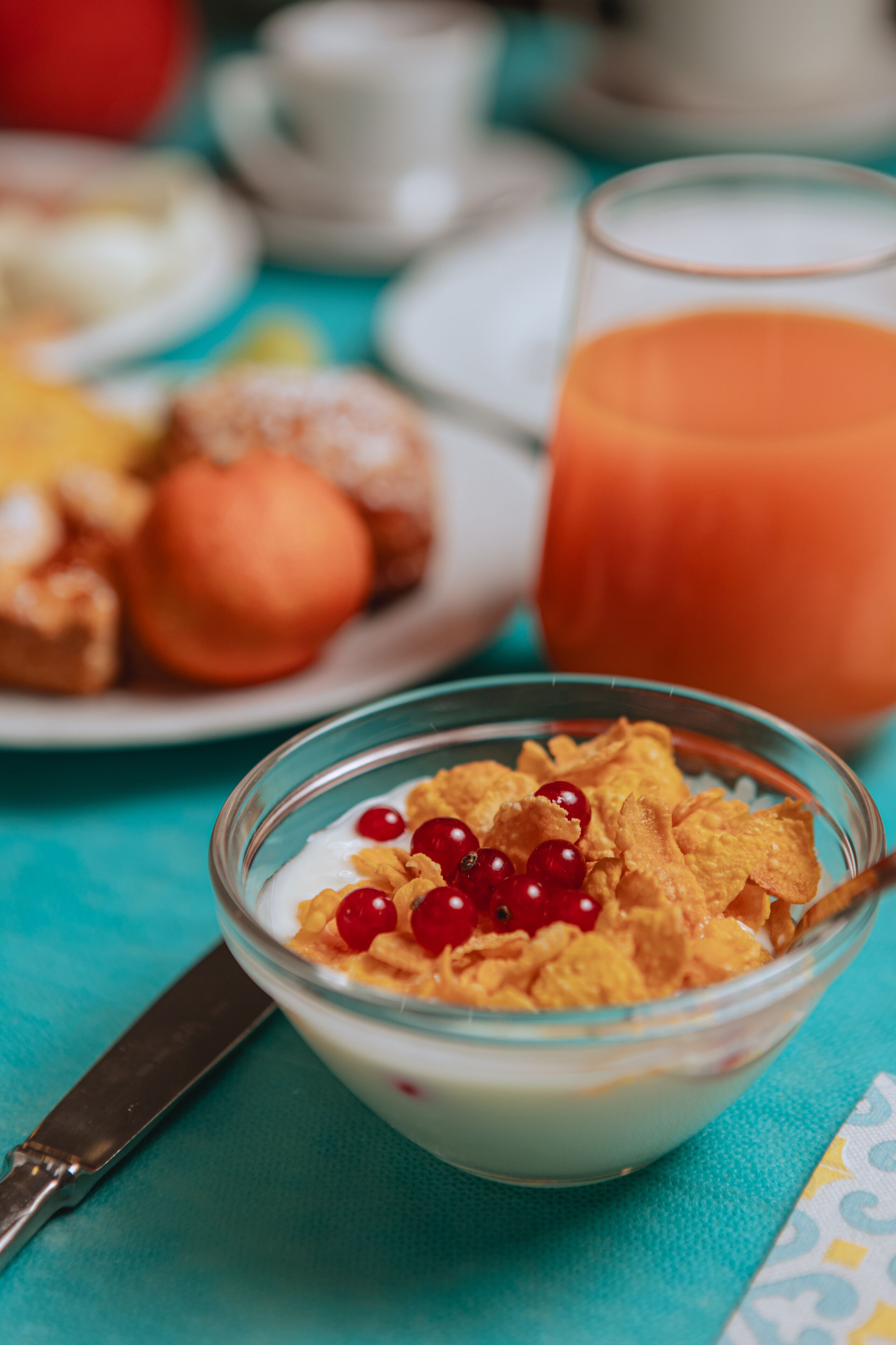 A bowl of cereal with milk and red berries, a glass of orange juice, and a plate with pastries on a turquoise table.