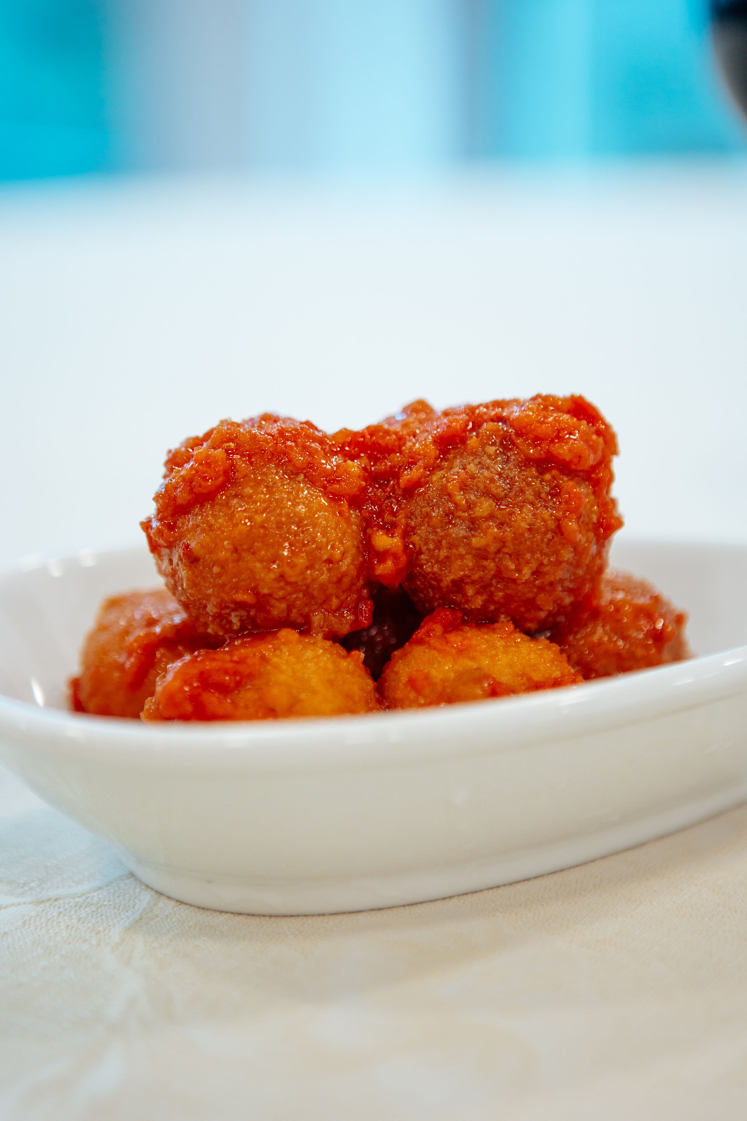 A white bowl containing several round, breaded, and fried items with a red sauce, placed on a white surface.