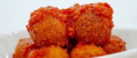 Un bol blanc contenant plusieurs aliments ronds, panés et frits, nappés d’une sauce rouge, le tout posé sur une surface blanche.