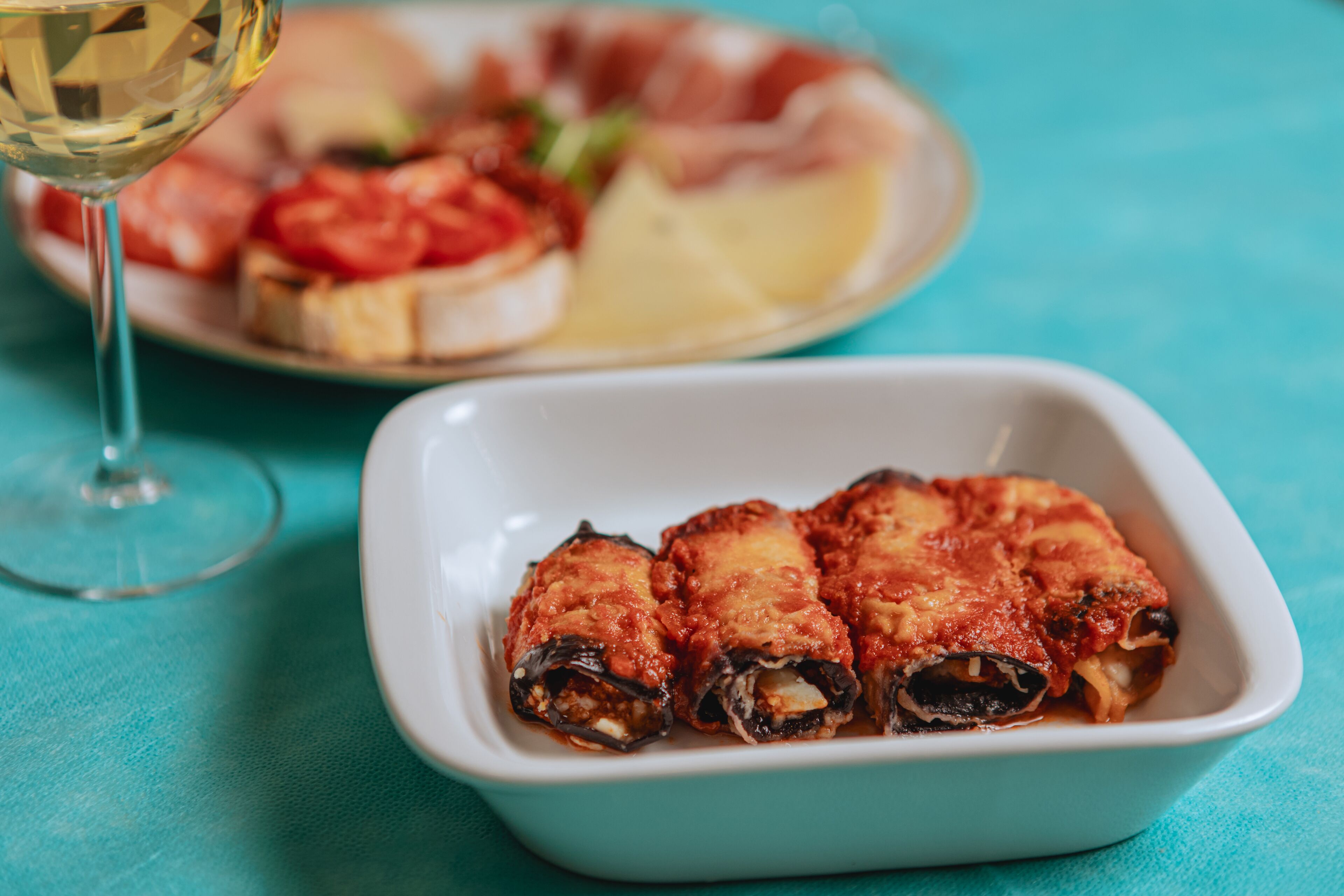 Eine weiße, rechteckige Schale mit Nudeln, rote Soße, ein Glas Weißwein und im Hintergrund ein Teller mit Tomatenscheiben und Käse.