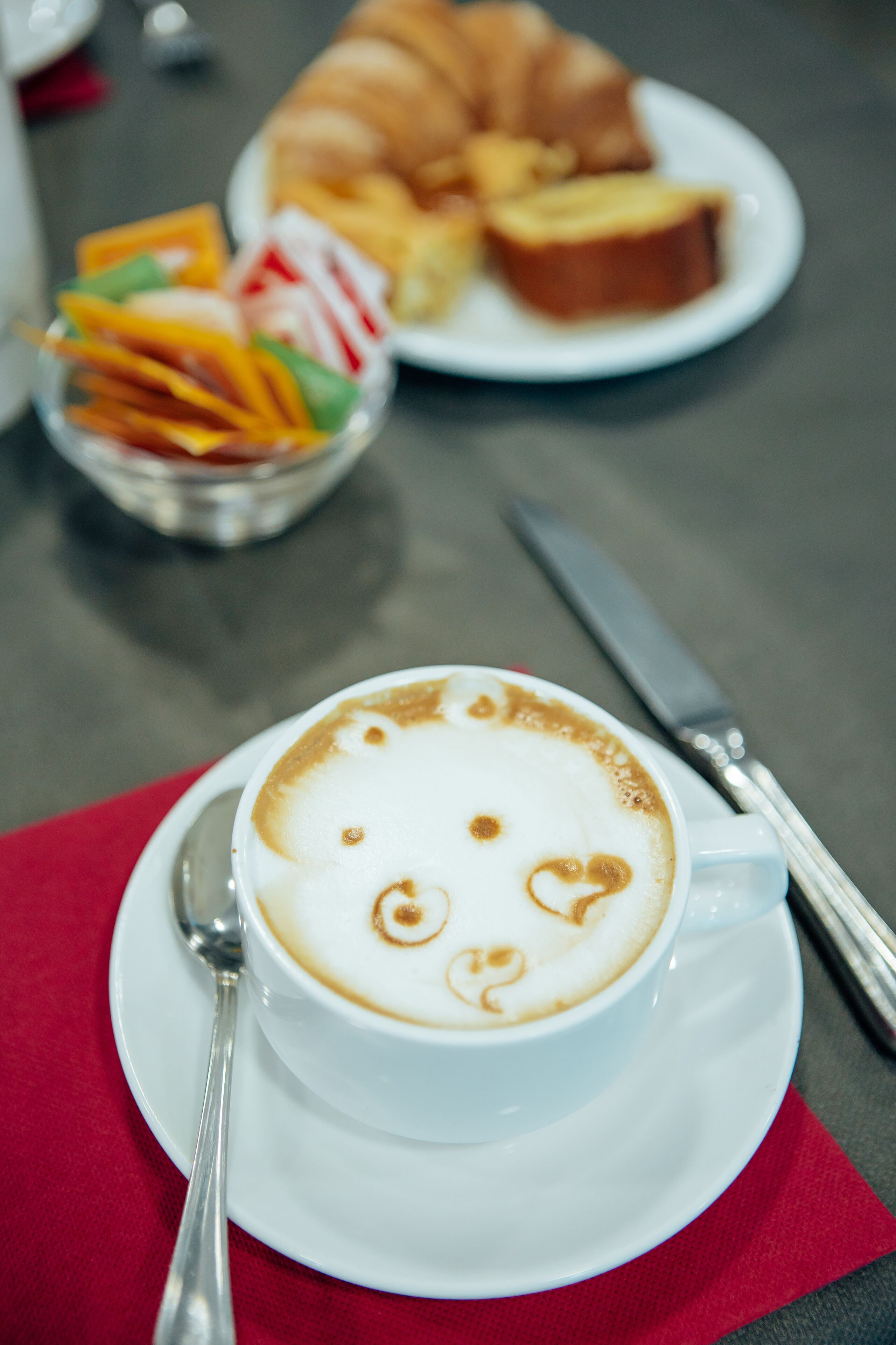 A cup of coffee with latte art on a saucer, a spoon, a knife, and a plate with food items in the background.
