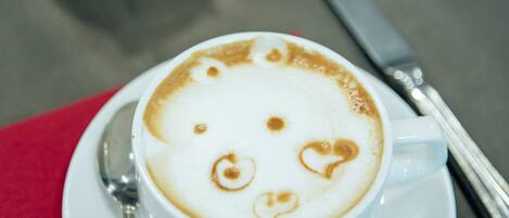 A cup of coffee with latte art on a saucer, a spoon, a knife, and a plate with food items in the background.
